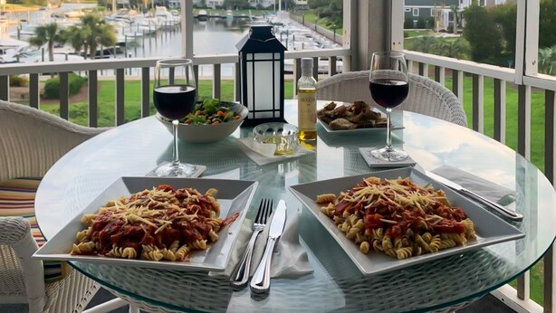 Une table à manger avec deux assiettes de pâtes, des verres à vin et une bouteille d’huile d’olive, le tout placé sur un balcon donnant sur une marina.