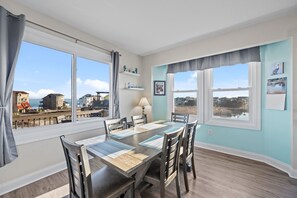 A dining area with a table set for six, large windows with curtains, a view of the beach, and a shelf with decorative items.