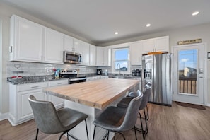 A modern kitchen with white cabinetry, a wooden island, stainless steel appliances, and a view of the city through the window.