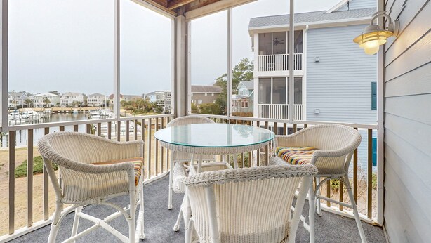 A covered balcony with wicker chairs and a glass table, overlooking a marina and residential buildings. - Mystic Marina B: Spacious condo with marina & Intercoastal views, screened-in balconies, W/D & AC (Southport)