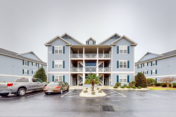 A multi-story residential building with a central balcony, parking lot, and a small palm tree.