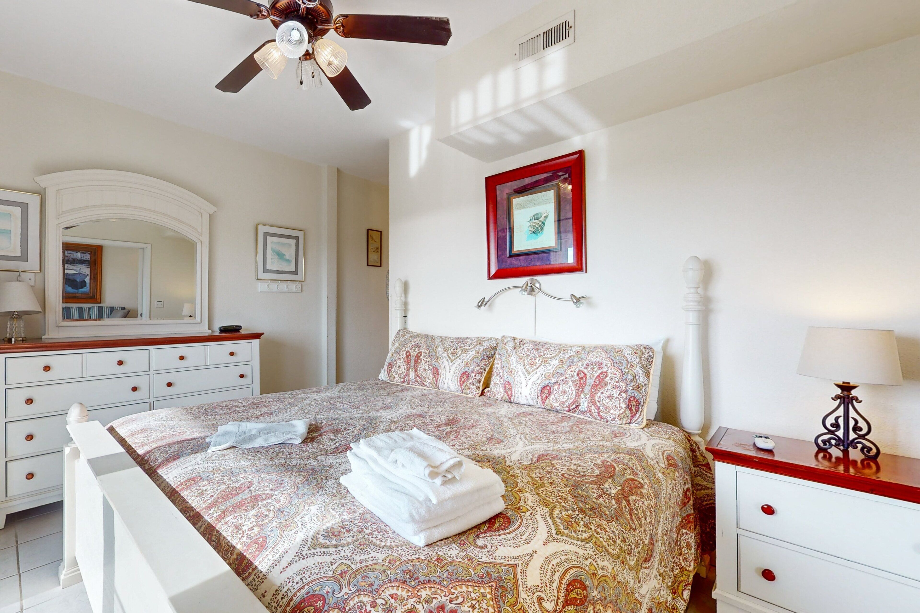 A bedroom with a bed, a nightstand, a dresser, a ceiling fan, and a framed picture on the wall.