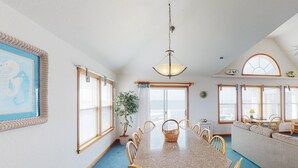 A dining room with a large table, chairs, a chandelier, and a view of the ocean through large windows.
