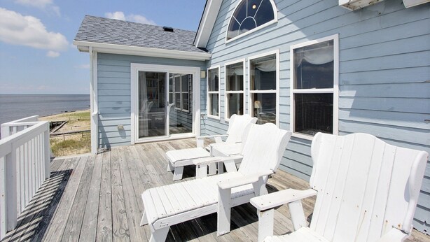 A coastal house with a deck featuring white Adirondack chairs and a glass door leading to the beach.
