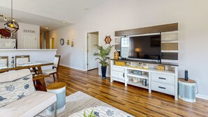 A modern living room with a wooden floor, a TV, a dining area with a white table and chairs, and a kitchen area with a fridge and cabinets. - Sunset Village 16-D: Top Floor, Pool, Hot Tub (Sunset Beach)