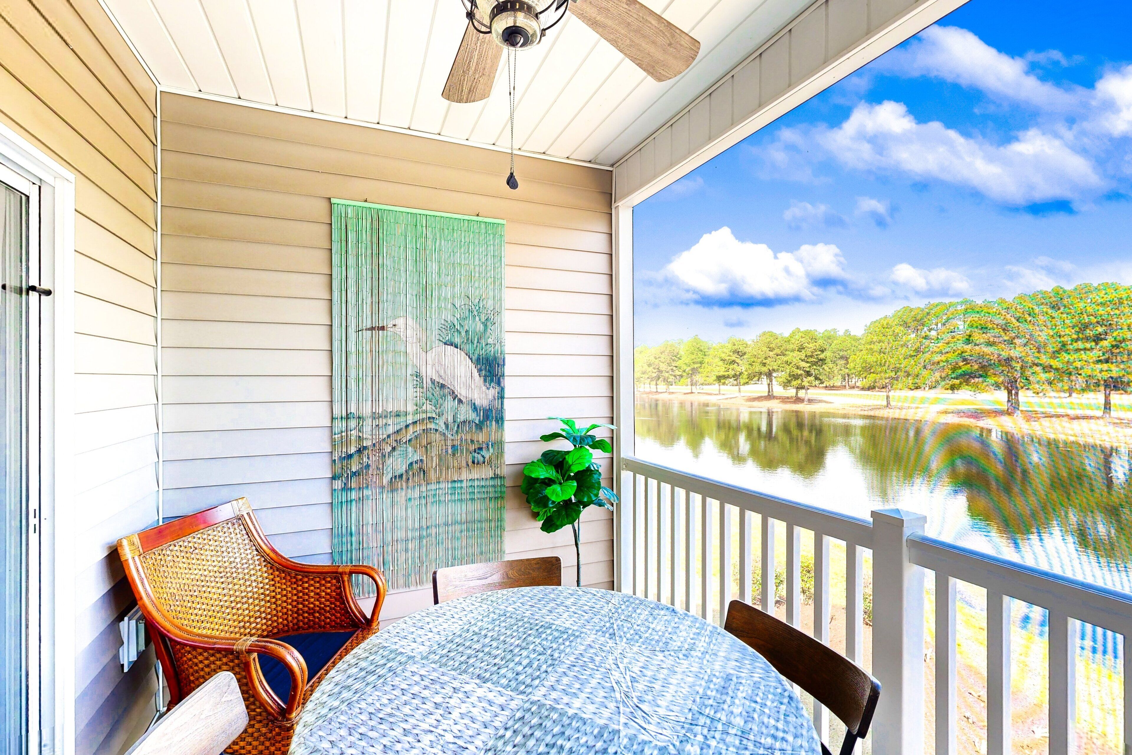 A covered outdoor area with a table and chairs, a ceiling fan, a large window overlooking a body of water and trees.