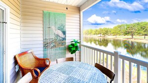 A covered outdoor area with a table and chairs, a ceiling fan, a large window overlooking a body of water and trees. - Sunset Village 16-D: Top Floor, Pool, Hot Tub (Sunset Beach)