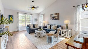 A spacious living room with hardwood flooring, a ceiling fan, a large window with curtains, a sofa set, a coffee table, and a side table with a lamp. - Sunset Village 16-D: Top Floor, Pool, Hot Tub (Sunset Beach)