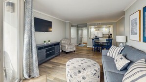Un soggiorno con un divano grigio, una poltrona a righe, un poggiapiedi con stampe nautiche, una TV a schermo piatto e un'area pranzo con sedie blu, con una cucina sullo sfondo.