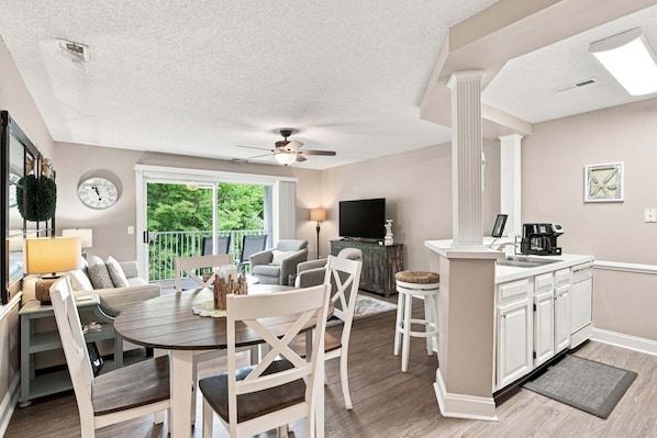 A modern kitchen with white cabinetry, a dining area with a round table and chairs, a ceiling fan, and a view of greenery outside.