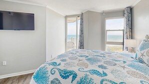 A bedroom with a bed, a TV mounted on the wall, a window with curtains, and a view of the sea. - Oceanfront retreat with pool, sauna, beach access, and golf nearby (North Topsail Beach)