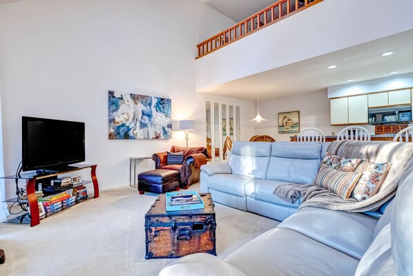 A living room with a TV, a sofa, a coffee table, and a bookshelf. - Golf Retreat at Club Villas: Cozy with Patio (Sunset Beach)