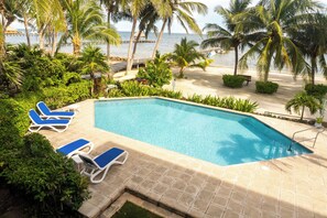 Una piscina rectangular con sillas reclinables azules y blancas, rodeada de palmeras y vista al océano.