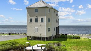 Uma casa de vários andares com telhado pontiagudo, erguida sobre pilares acima de um corpo d'água, com uma área gramada em primeiro plano e uma vista clara da água e do céu ao fundo.