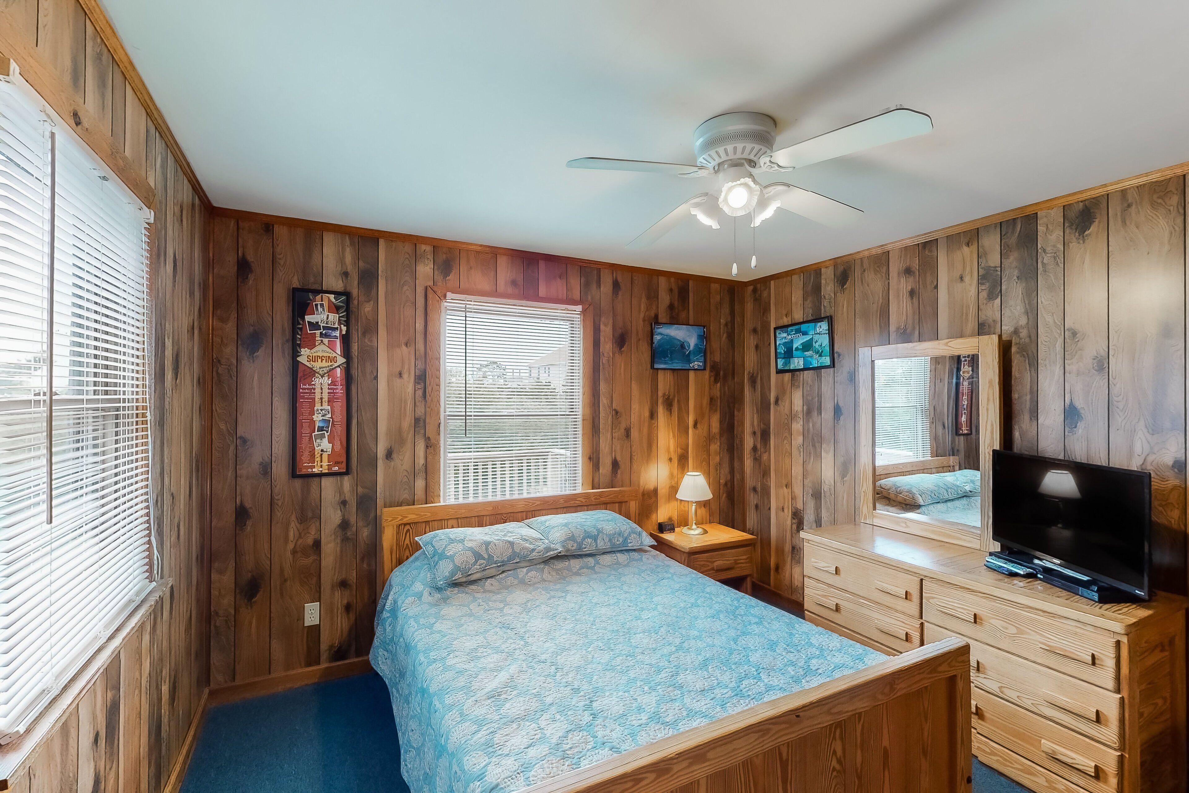A bedroom with wooden walls, a bed with a patterned bedspread, a ceiling fan, a TV on a dresser, and a window with blinds.