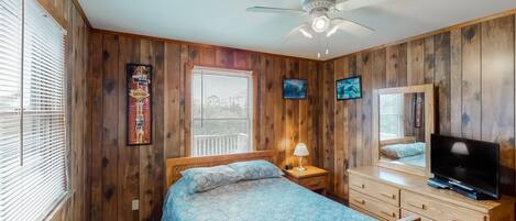A bedroom with wooden walls, a bed with a patterned bedspread, a ceiling fan, a TV on a dresser, and a window with blinds.