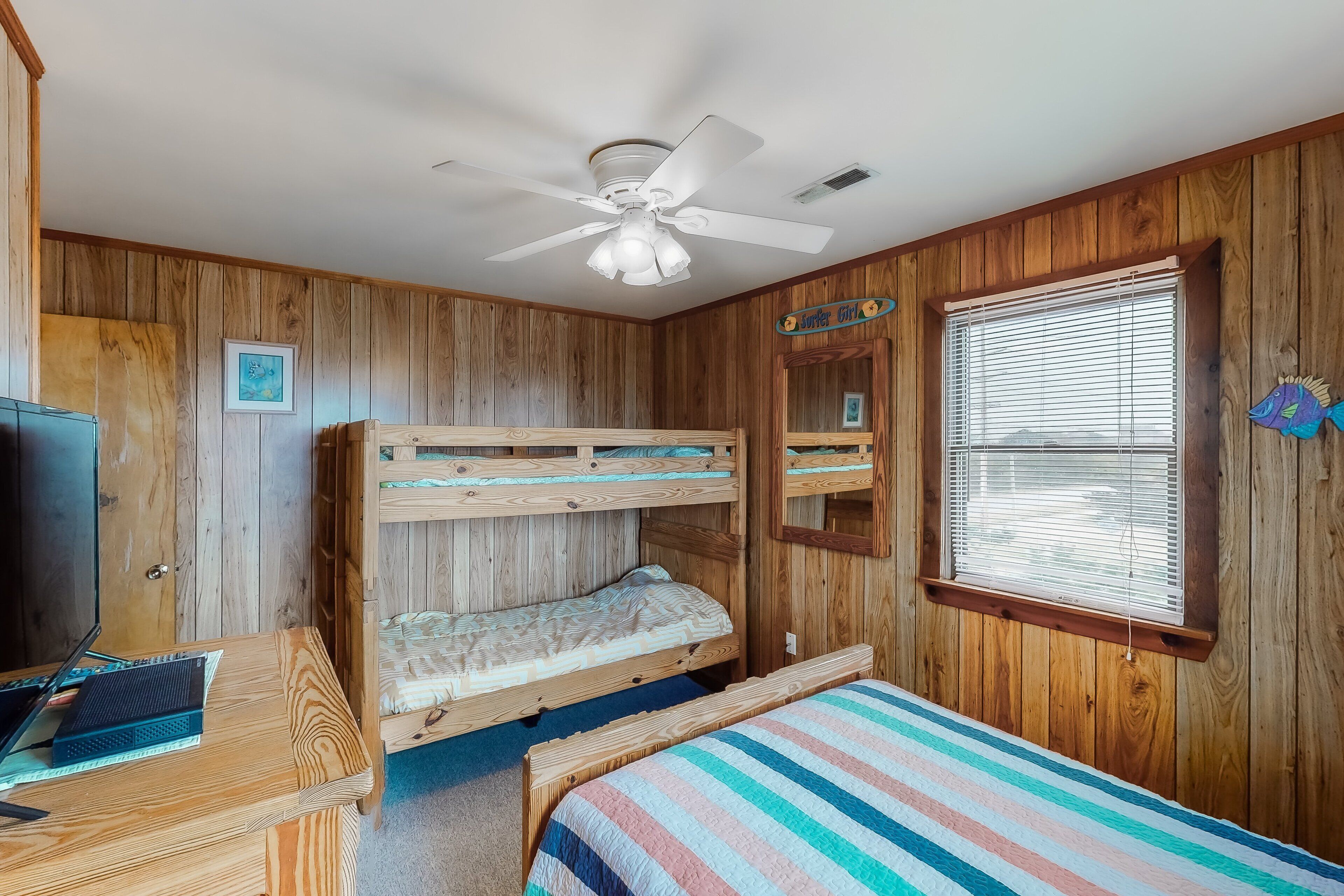 A room with wooden walls, a bunk bed, a desk with a laptop, a ceiling fan, and a window with blinds.