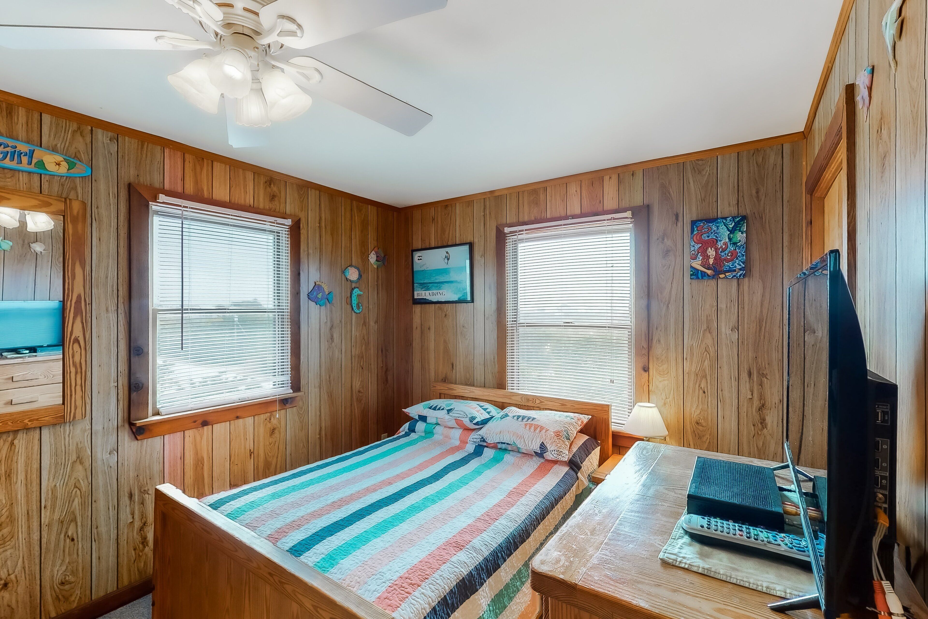 A bedroom with wooden walls, a bed with a striped blanket, a desk with a computer, and a ceiling fan.