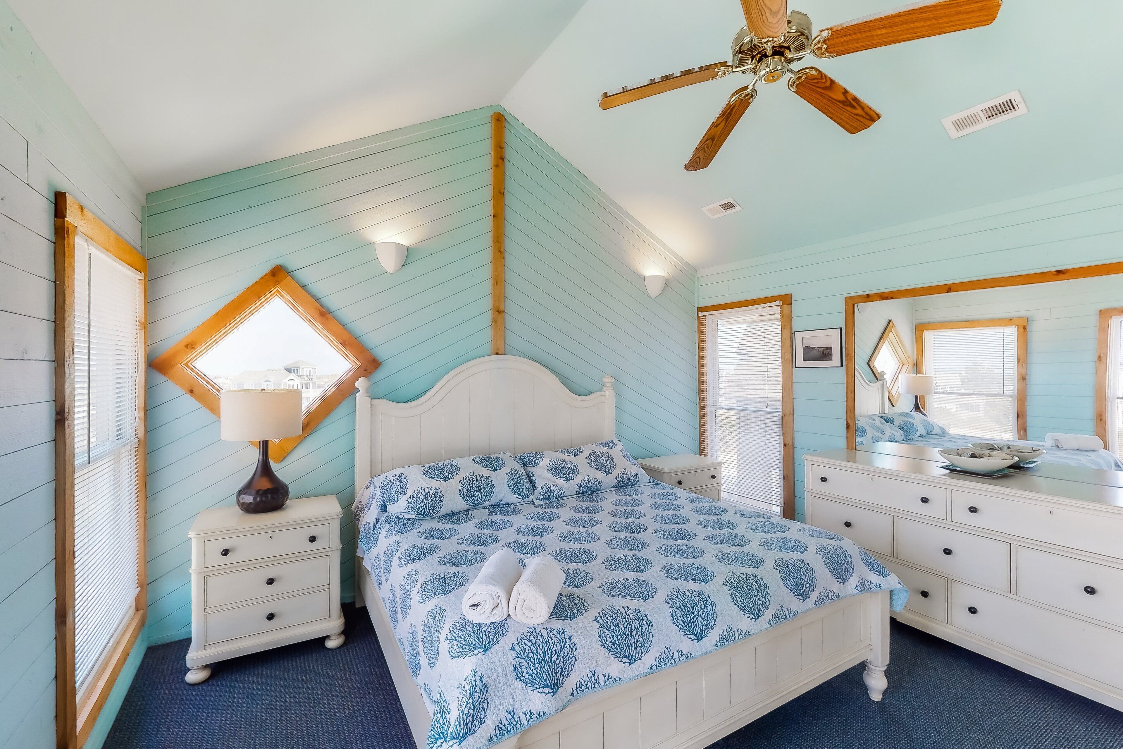 Une chambre à coucher comprenant un lit, des tables de chevet, une commode, un ventilateur de plafond et une fenêtre avec des stores.