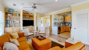 A spacious living room with a large orange sofa, a coffee table, and a TV. There is a kitchen area with wooden cabinets and a dining area with a small table and chairs. - Vibrant beach retreat with washer/dryer, central AC, deck, & pool access (Hatteras)