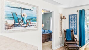 A living room with a beach-themed painting, a wooden bench, and a view into a bedroom with a bed and a nightstand.