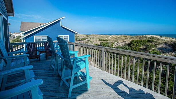 Um deck de madeira com cadeiras Adirondack azuis e vista para a praia.