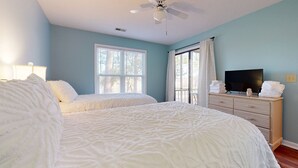 Une chambre avec deux lits, un ventilateur de plafond, un téléviseur posé sur une commode et une fenêtre avec des rideaux.