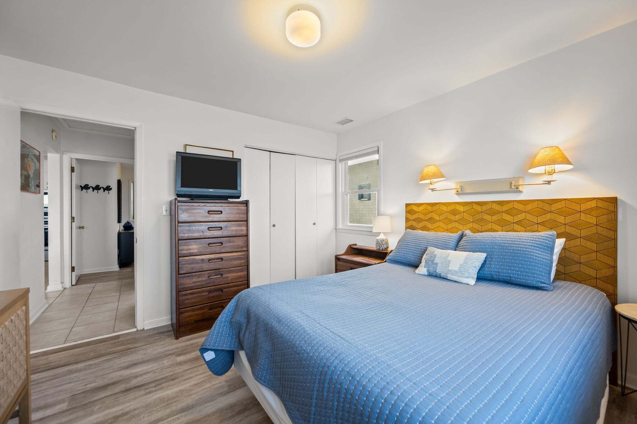 A bedroom with a bed, a TV, a dresser, a nightstand, and wall-mounted lamps.