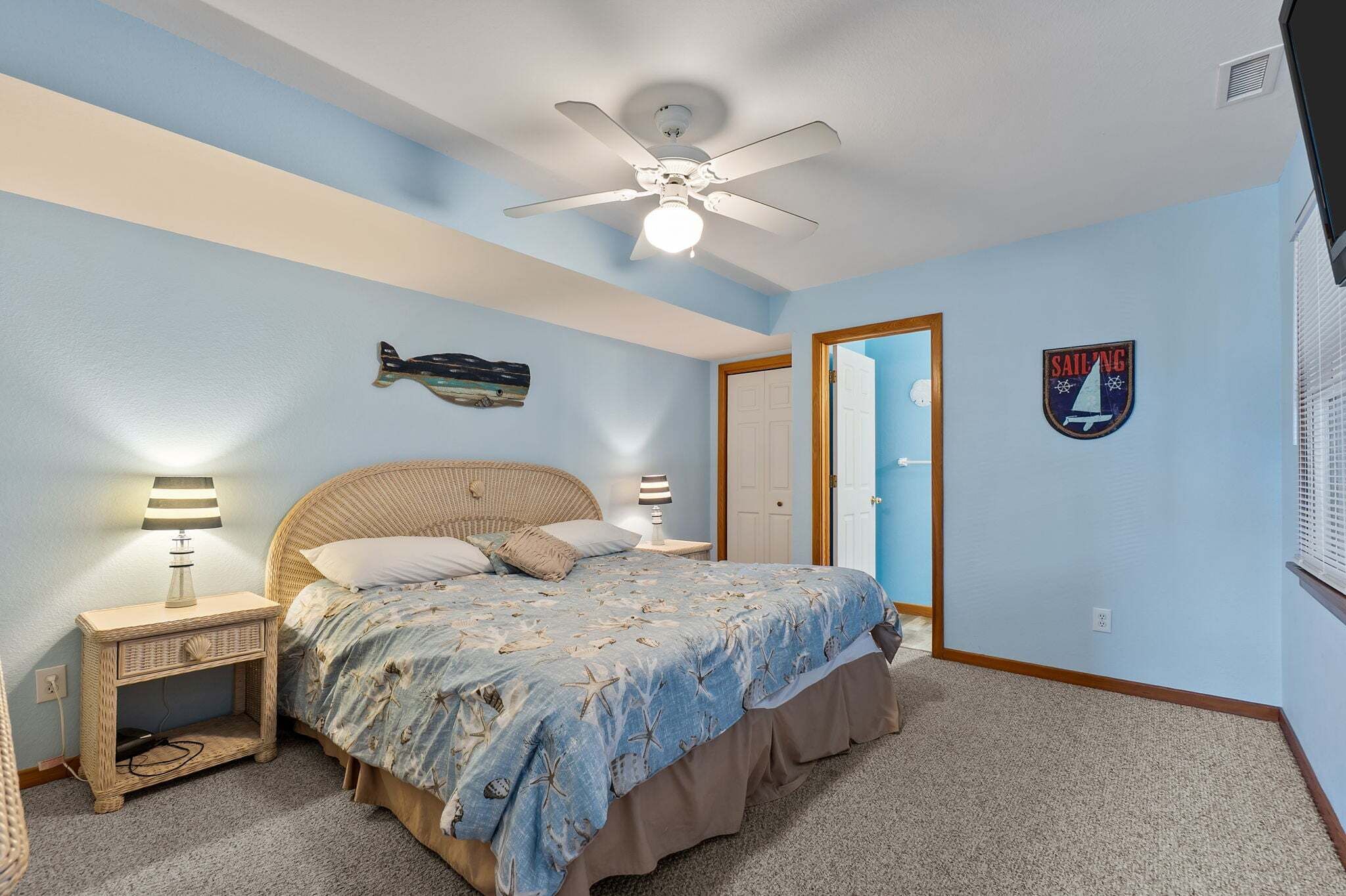 A bedroom with a bed, a ceiling fan, a wall-mounted fish decoration, a sailboat sign, and a door.