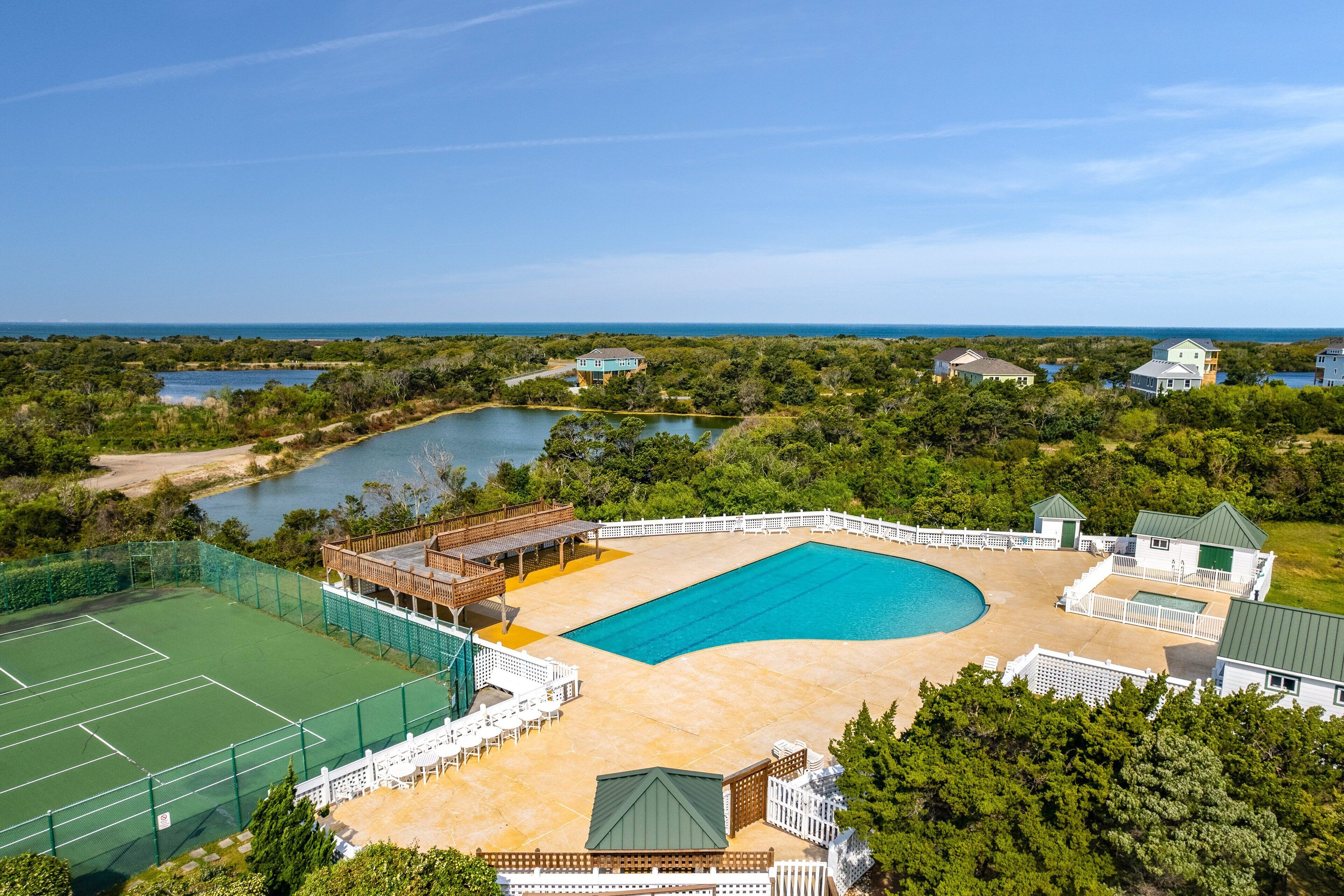 Ein großer Swimmingpool, umgeben von einer Holzterrasse und Tennisplätzen, mit Blick auf den Ozean und die umliegende Bepflanzung.