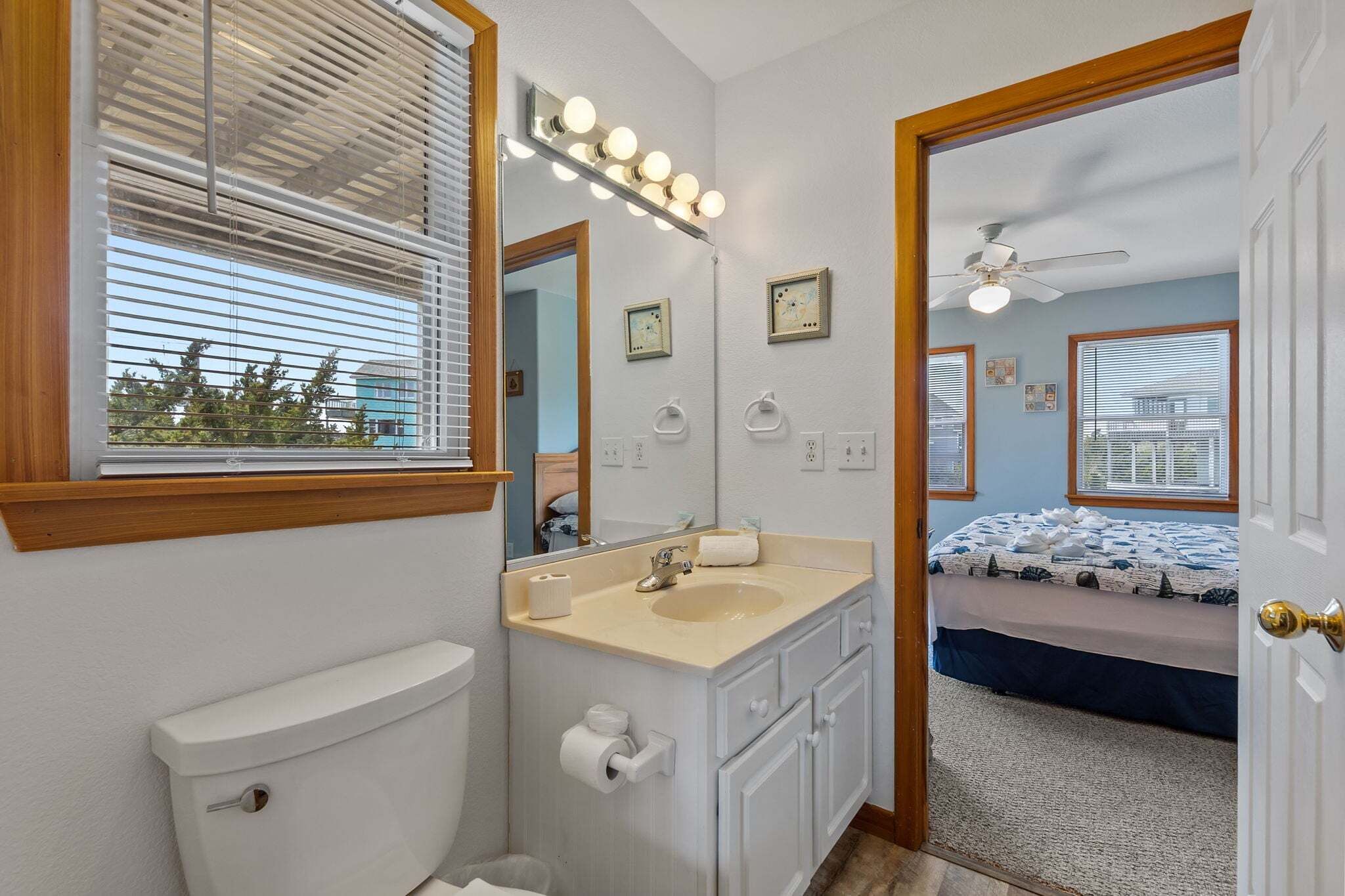 A bathroom with a toilet, sink, vanity, and a mirror with lights. There is a bed with a blue and white bedspread in the adjacent room.