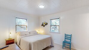 A bedroom with a bed, two windows, a chair, a nightstand, and a wall decoration.