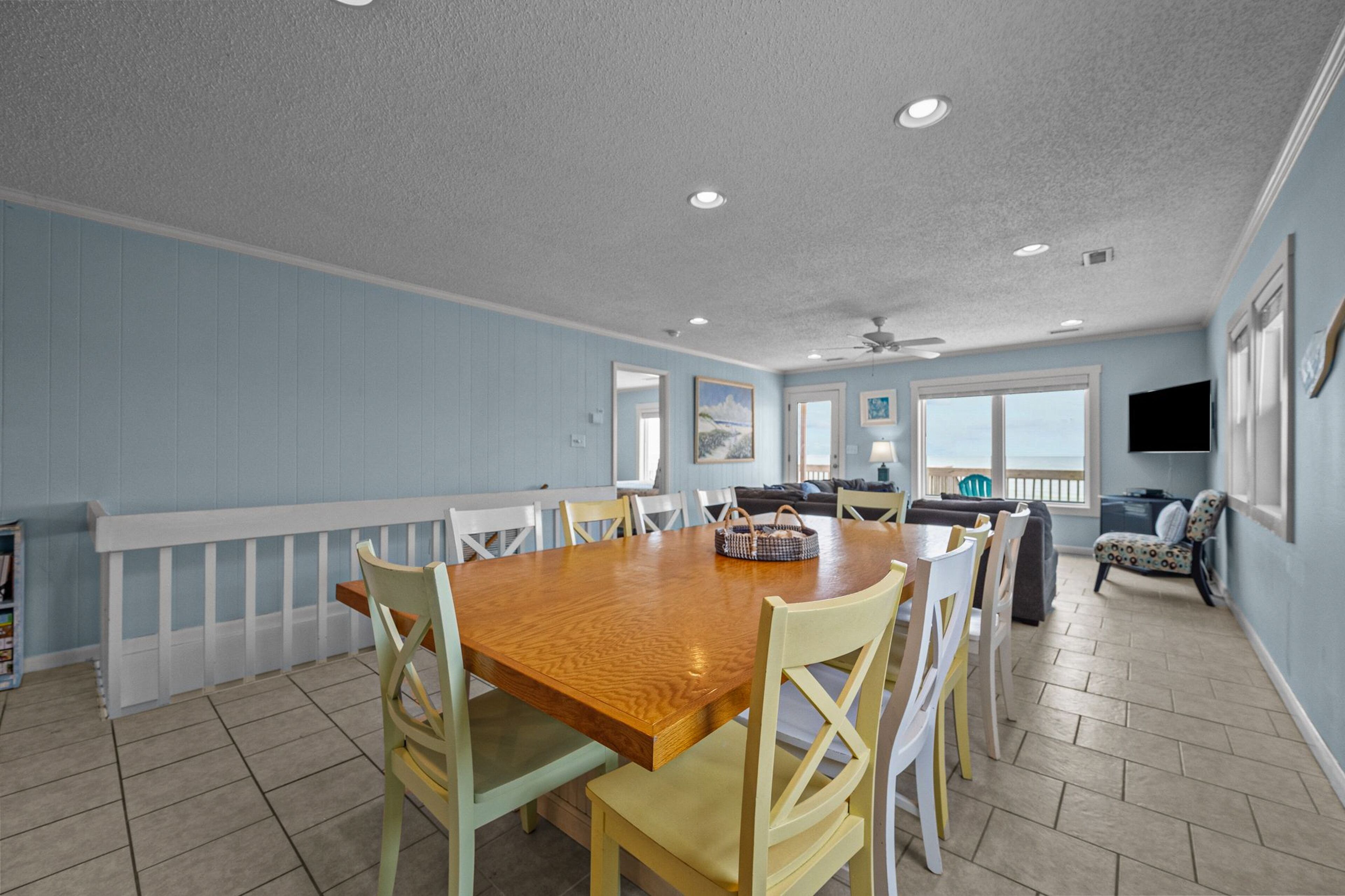A dining area with a wooden table and chairs, a ceiling fan, and a view of the ocean through large windows.
