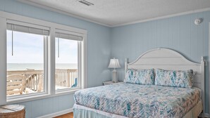 A bedroom with a large bed, a nightstand, a lamp, a wicker chair, and a view of the beach through large windows. - Oceanfront home with private beach access & outdoor shower - dog-friendly (Surf City)