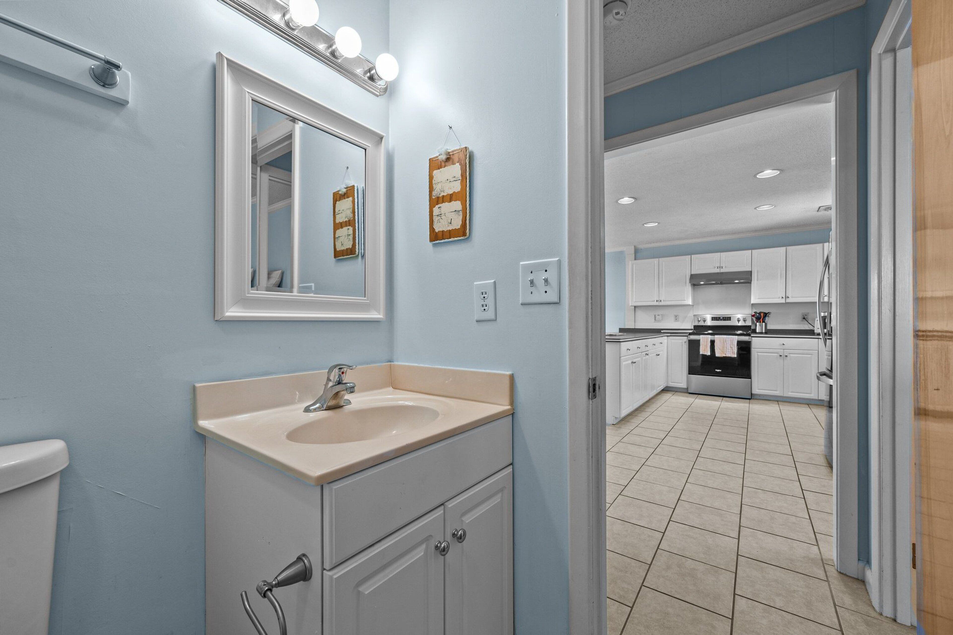 A bathroom with a sink, mirror, and lights. It leads to a kitchen with cabinets and appliances.