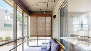 Een balkon met een zithoek met kussens, een glazen tafel en uitzicht op omliggende huizen.