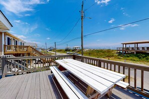 Een houten terras met picknicktafels, een trap naar een gebouw en een strand op de achtergrond.