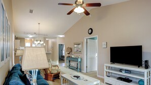 A living room with a ceiling fan, a television on a white stand, a sofa, a coffee table, and a lamp. - Coral Hideaway: Balcony | Pool | Dog Friendly (Sunset Beach)