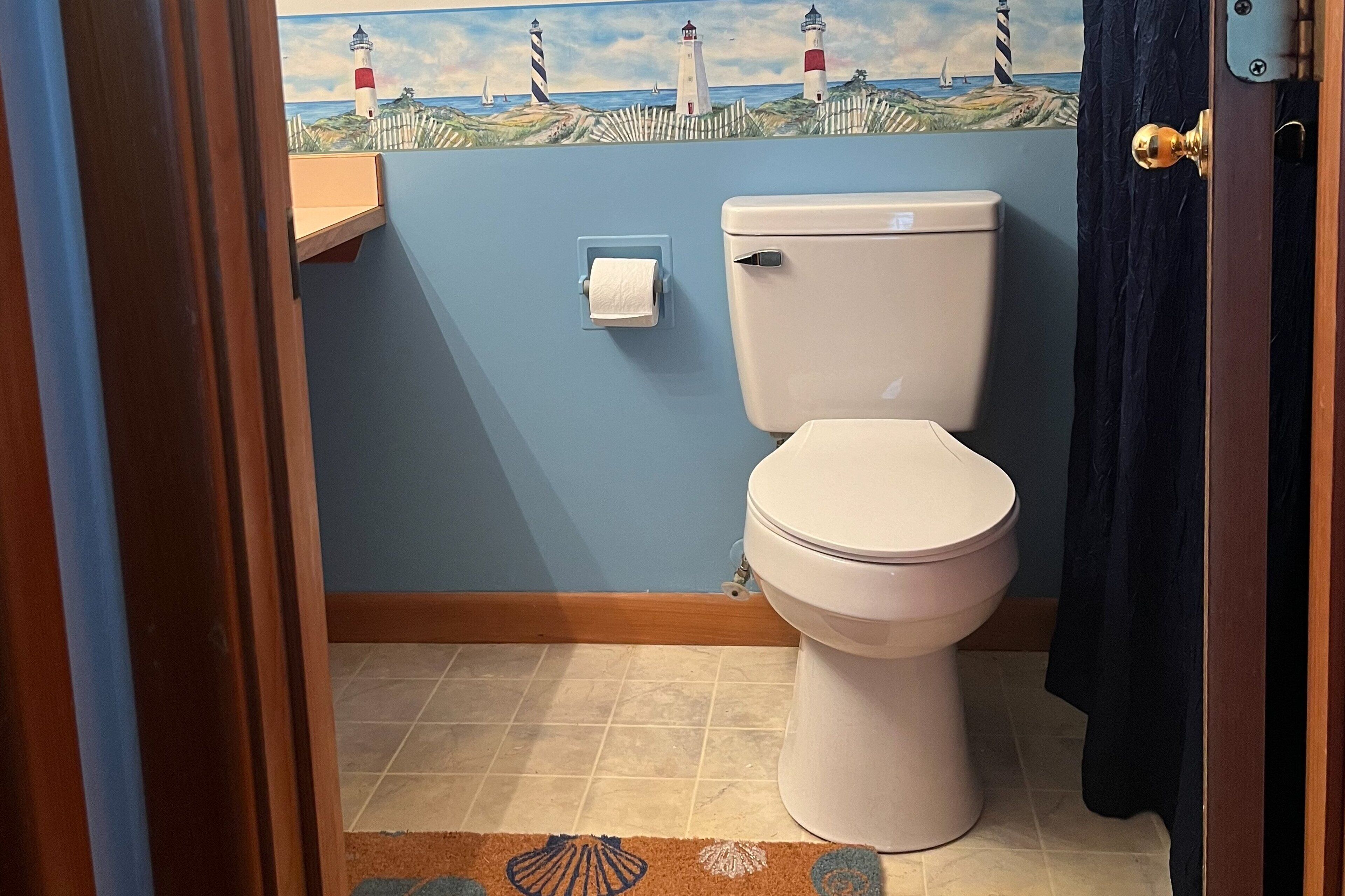 A bathroom with a toilet, a roll of toilet paper, a blue wall, a wooden shelf, and a decorative painting of lighthouses.