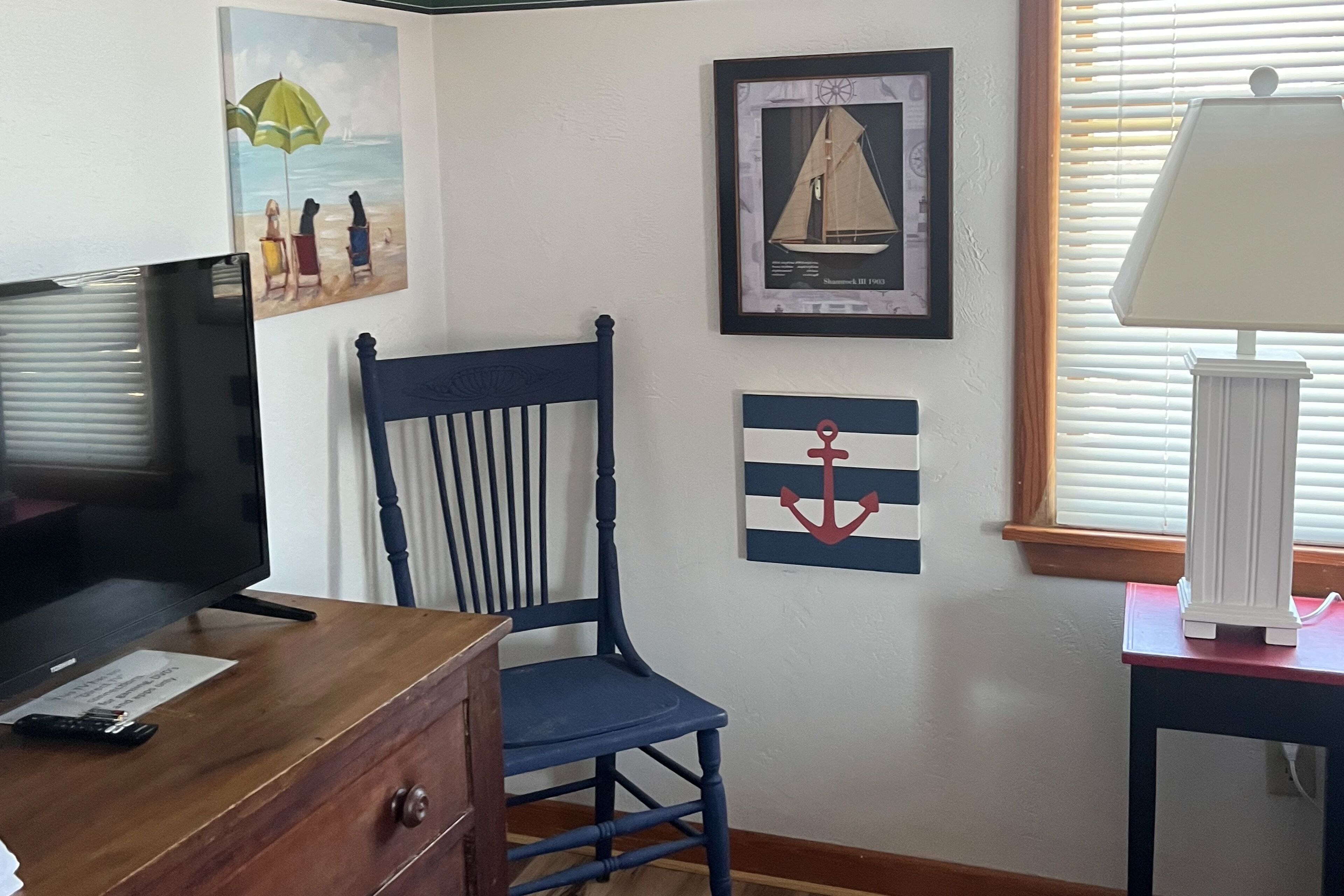 A room with a wooden dresser, a television, a blue chair, a framed sailboat painting, and a wall with an anchor design.