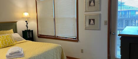 Un dormitorio con cama, mesita de noche, lámpara, dos ventanas con persianas y dos cuadros en la pared.