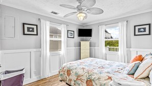 A bedroom with a bed, a ceiling fan, a TV, and a window with curtains. - Home near the beach with balconies, outdoor shower, grill, beach gear, W/D, & AC (Sunset Beach)