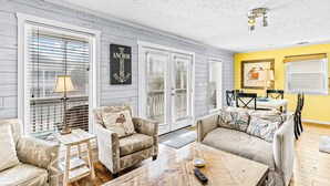 A living room with a wooden floor, two sofas, a coffee table, and a dining area with chairs and a table. - Home near the beach with balconies, outdoor shower, grill, beach gear, W/D, & AC (Sunset Beach)