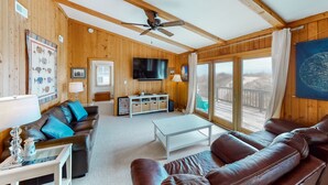 A wooden-paneled living room with a ceiling fan, a TV, a sofa, a coffee table, and a view through large windows. - Oceanfront, Dog-Friendly House w/ Free WiFi, Central AC, Private W/D, Ocean View (Hatteras)