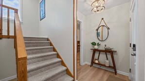 Un escalier recouvert de moquette avec des rampes en bois, un miroir fixé au mur, un luminaire suspendu et une petite étagère ornée d’objets décoratifs.