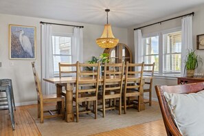 Uma área de refeições com mesa e cadeiras de madeira, luminária pendente e uma janela grande com cortinas brancas.