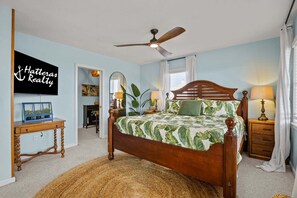 Quarto com cama, ventilador de teto, mesa de cabeceira, luminária, espelho e televisão fixada na parede.