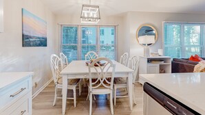 A dining area with a white table and chairs, a chandelier, and a view through the window blinds. - Ev's Escape: Sunroom, dog-friendly (Sunset Beach)