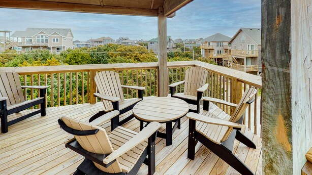 A wooden deck with Adirondack chairs and a round table, overlooking a residential area with houses and greenery. - Lovely home with private pool, decks, amazing ocean view, & W/D (Hatteras)