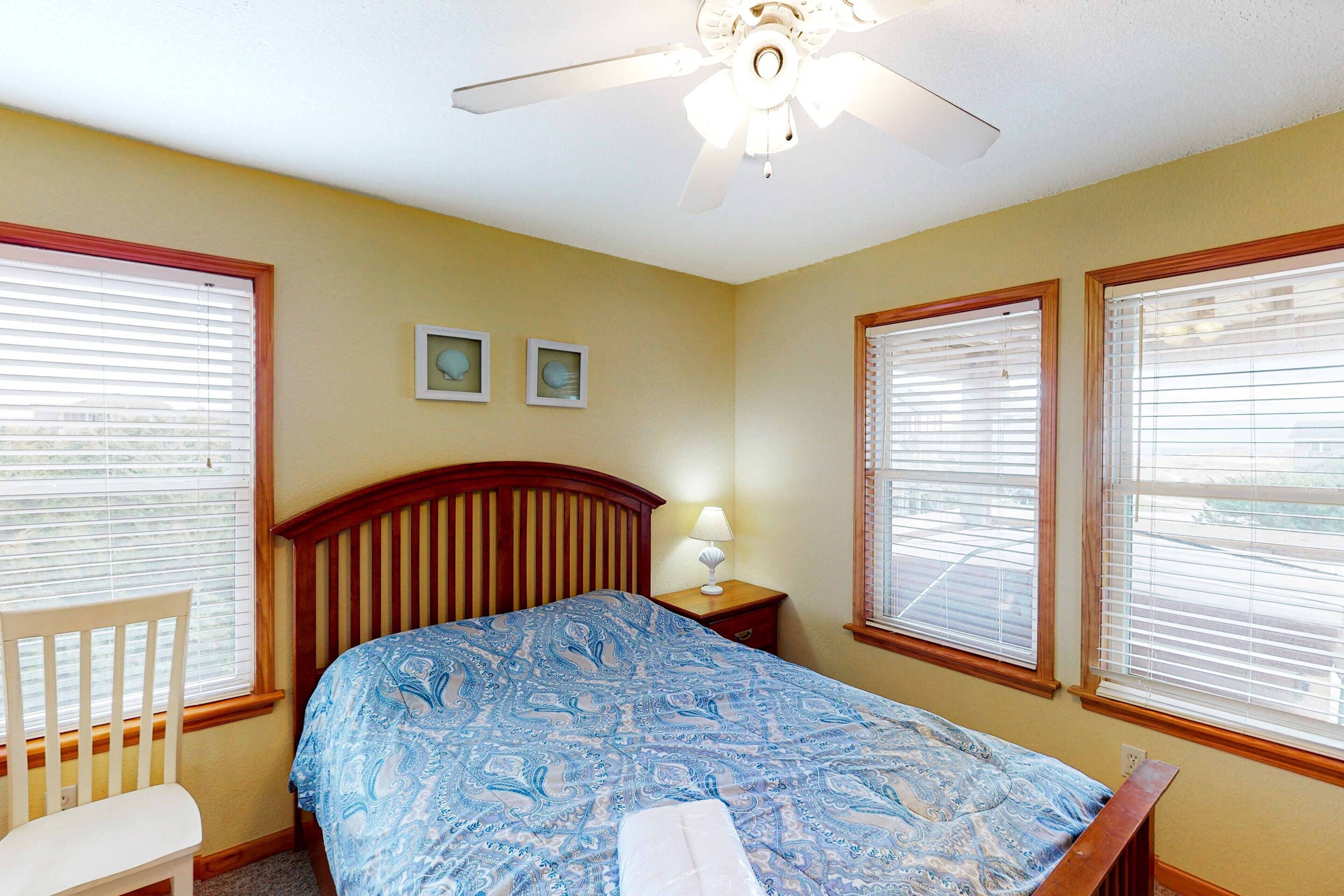 A bedroom with a bed, a chair, a nightstand, two wall-mounted pictures, and a ceiling fan.